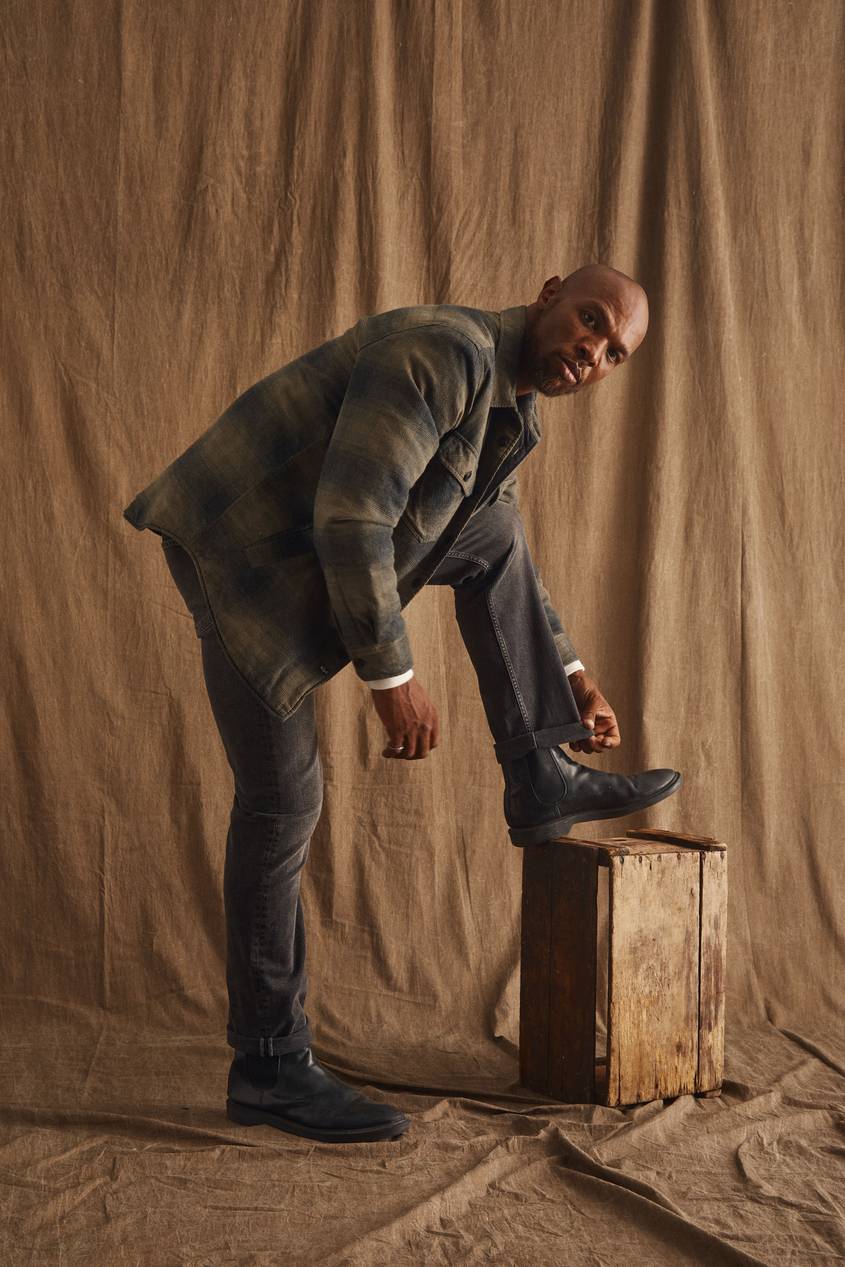 A man with a short beard leans forward against a warm brown fabric backdrop, tying the laces of a black leather boot with his foot propped on a wooden crate. He wears a plaid shacket in muted green and gray tones over dark slim-fit jeans and a black leather belt