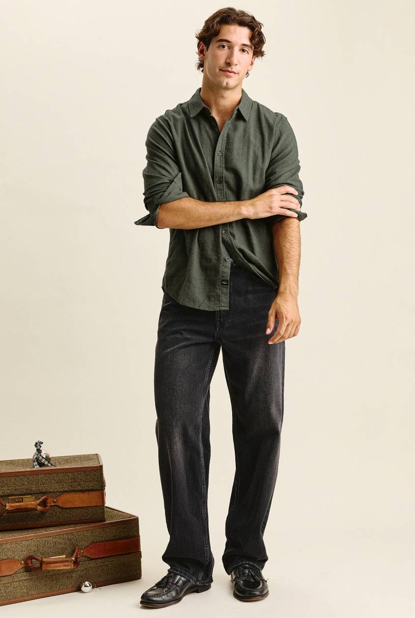 Man with wavy brown hair standing with one hand rolling up his sleeve, wearing an olive green button-up shirt, wide-leg faded black jeans, and black tassel loafers. Vintage tweed suitcases with leather straps are stacked beside him.