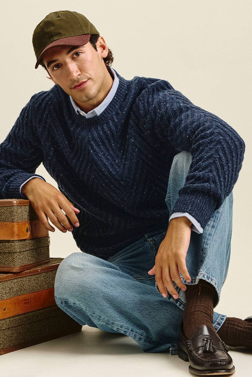 Man seated on the floor in an olive and burgundy baseball cap, a navy speckled chevron-knit sweater over a light blue collared shirt, light-wash jeans, brown ribbed socks, and dark tassel loafers. He leans on a stack of tweed suitcases.