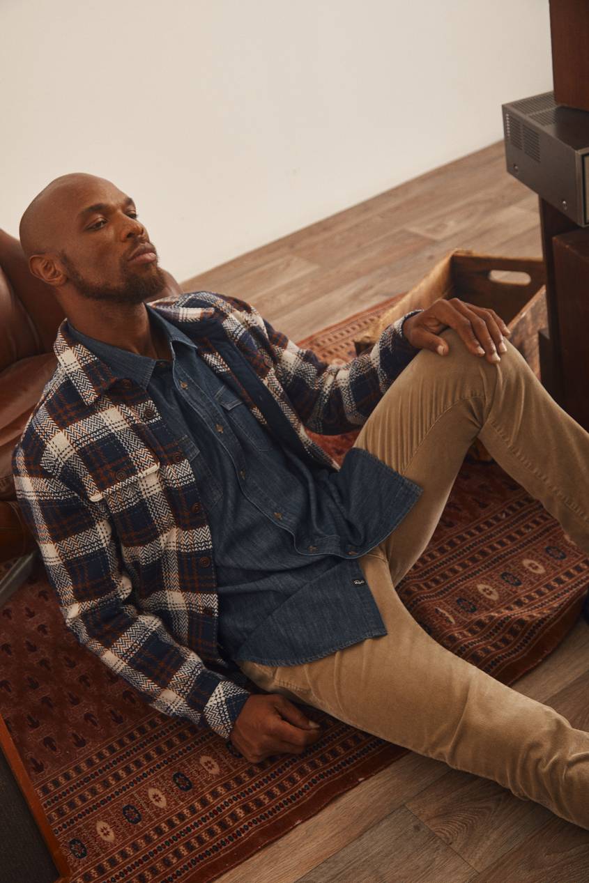 Man sitting while wearing a plaid overshirt, green button down, and brown pants.
