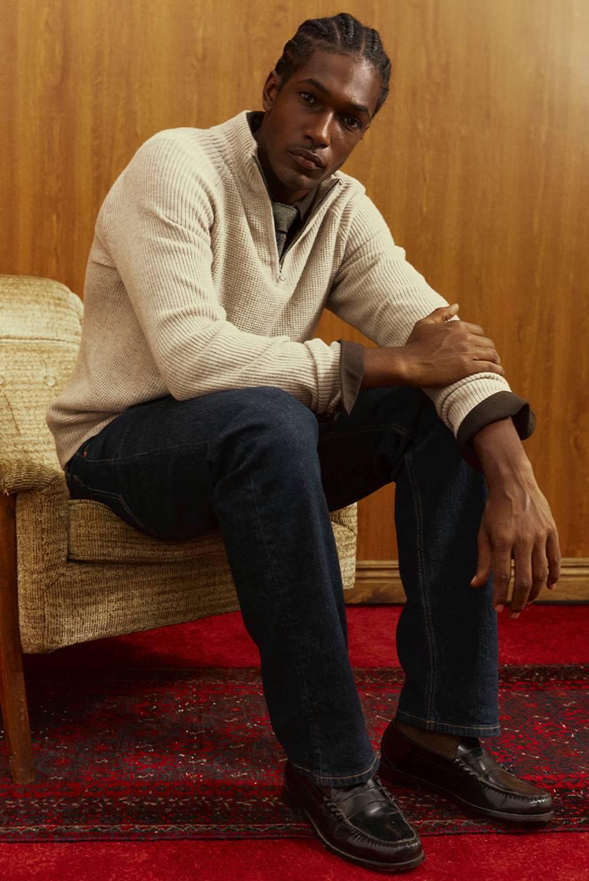 Man with braided hair seated in a tan upholstered chair, wearing a cream waffle-knit quarter-zip sweater over a collared shirt, dark-wash jeans, and burgundy penny loafers. He sits on a red patterned rug against a wood-paneled wall.