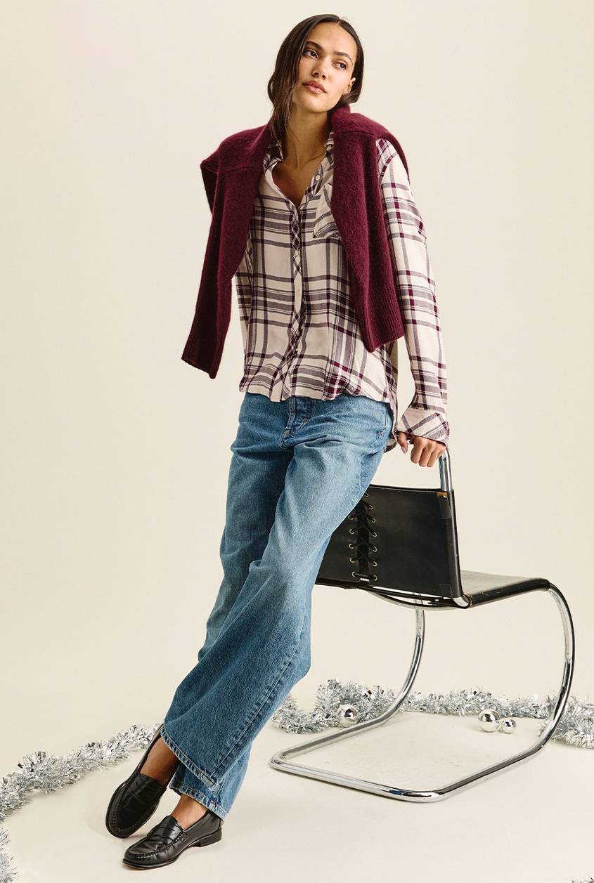 Woman leaning on a black and chrome chair, wearing a cream and burgundy plaid button-up shirt with a burgundy sweater draped over her shoulders, light-wash wide-leg jeans, and black loafers. Silver tinsel and ornaments are scattered on the floor.