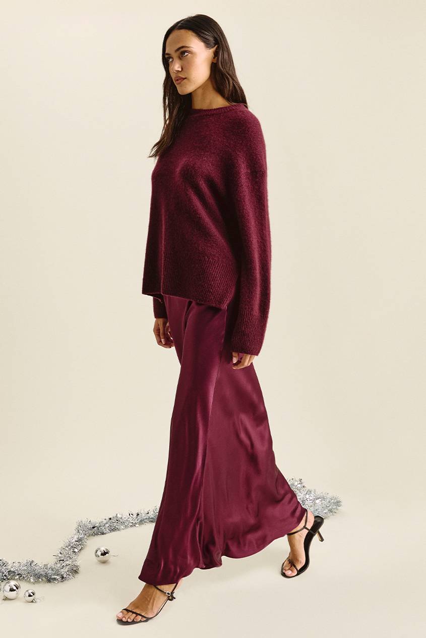 Woman walking and looking to the side, wearing an oversized burgundy sweater over a matching burgundy satin maxi skirt, with black strappy heels. Silver tinsel and ornaments lie on the floor.