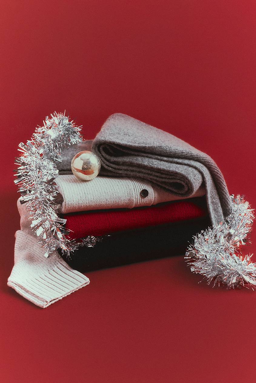 Stack of folded knitwear on a red background, with a gray scarf, a cream cardigan, a red sweater and a black sweater, decorated with silver tinsel and a gold ornament.