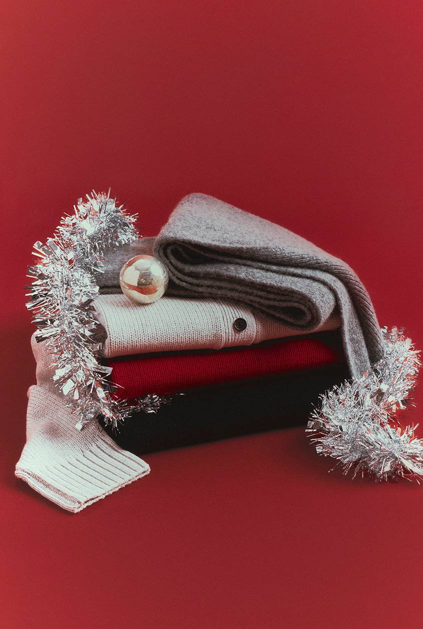 Stack of folded knitwear on a red background, with a gray scarf, a cream cardigan, a red sweater and a black sweater, decorated with silver tinsel and a gold ornament.