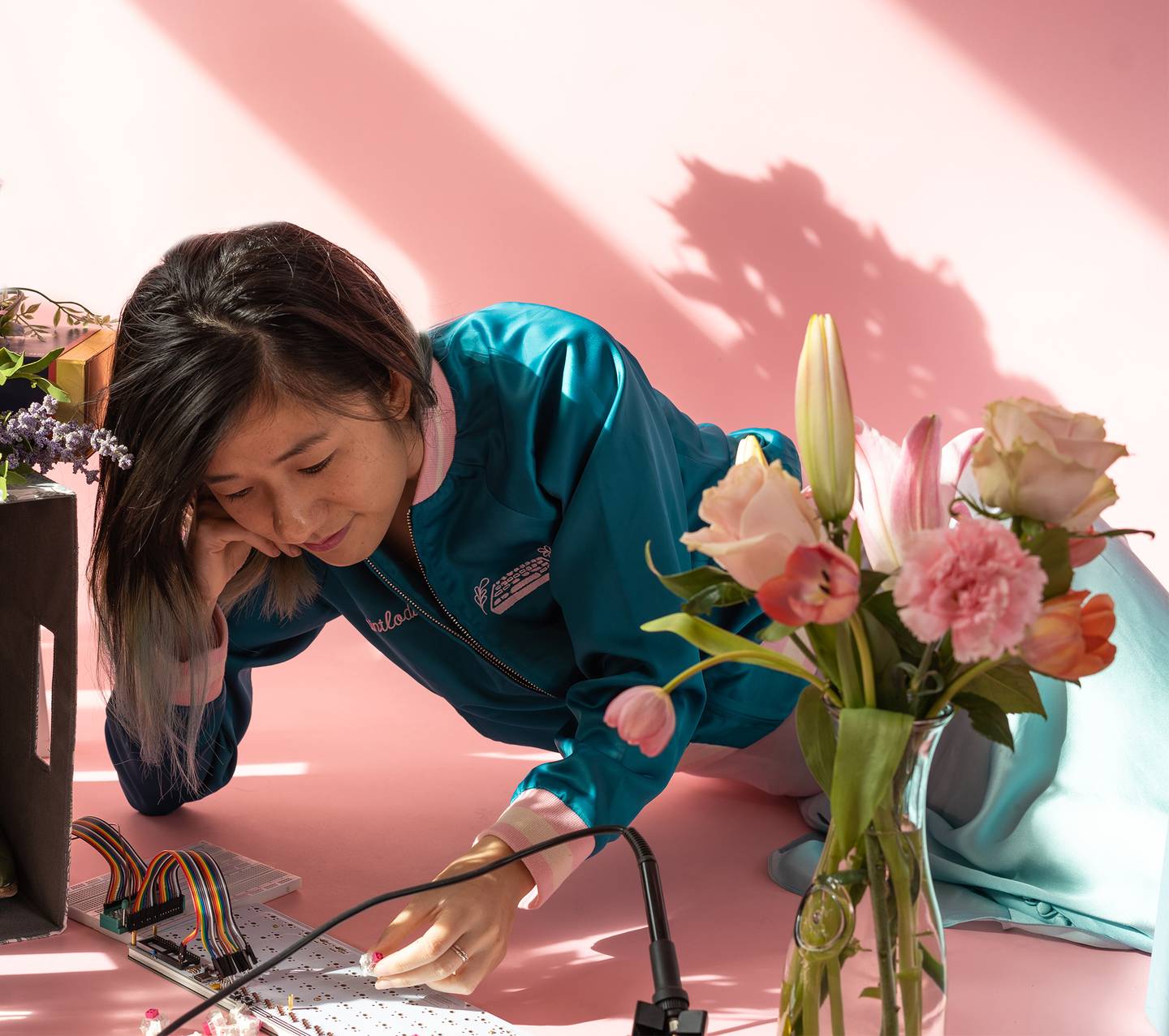 Photo of Susan Lin who is the Owner, Founder, Creator of Magic Girl and Mintlodica in a pink room with flowers, soldering tools, and a mechanical keyboard PCB.