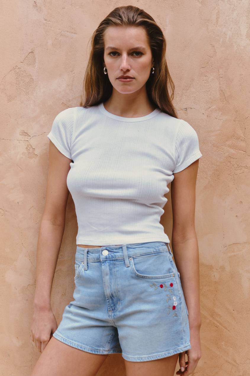 Woman standing against a textured wall wearing a white ribbed short-sleeve top and light-wash denim shorts with embroidered floral details on the front.