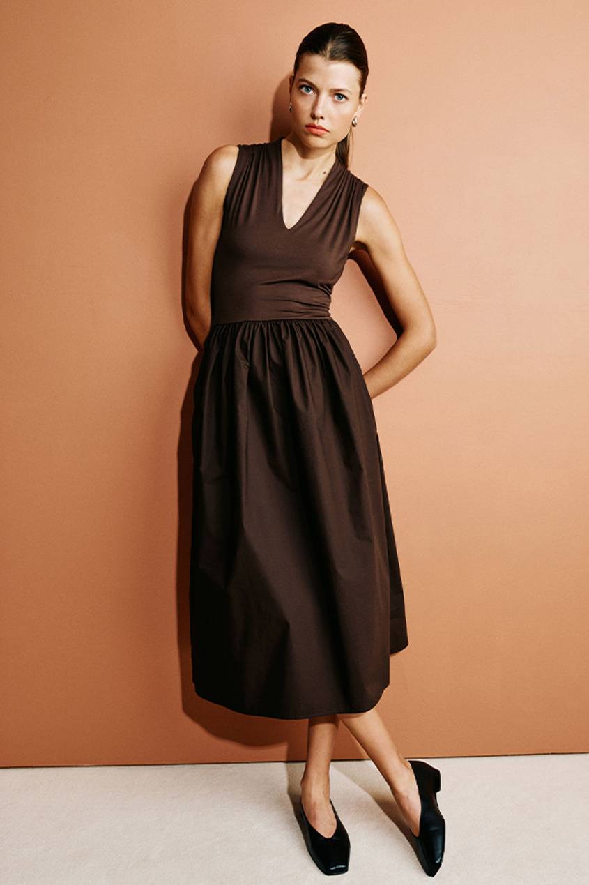 Woman posing against an orange background while wearing a brown midi dress and black ballet flats.