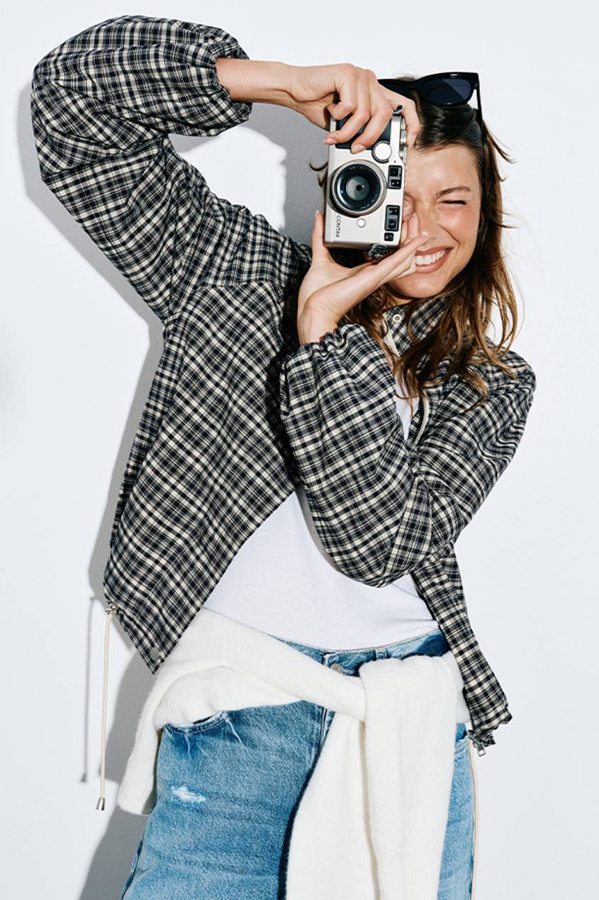 Woman pretending to take a picture while wearing a plaid checkered jacket, white shirt, and blue jeans.