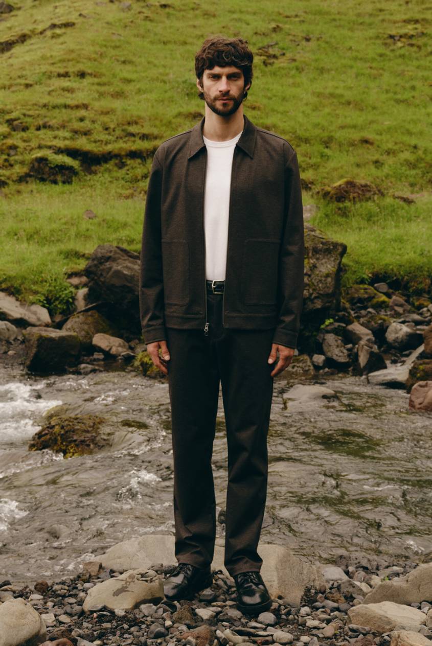 Model wearing a dark brown jacket over a white T-shirt with black pants, standing in a rocky outdoor setting.