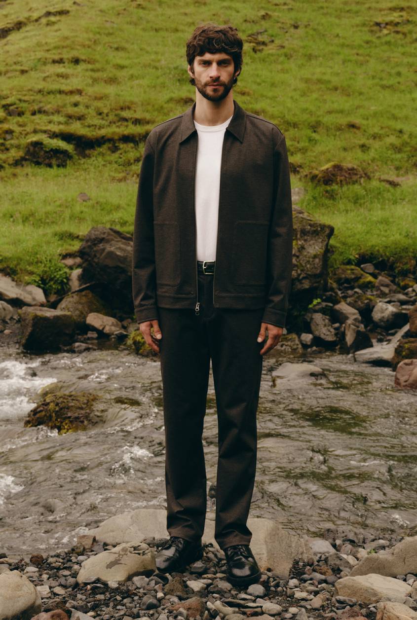 Model wearing a dark brown jacket over a white T-shirt with black pants, standing in a rocky outdoor setting.