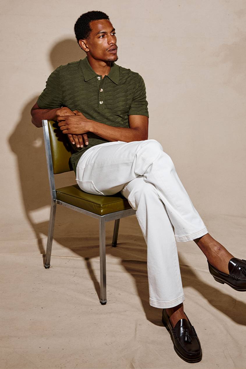 Man wearing a green polo with cream pants while sitting in a green chair against a beige background.