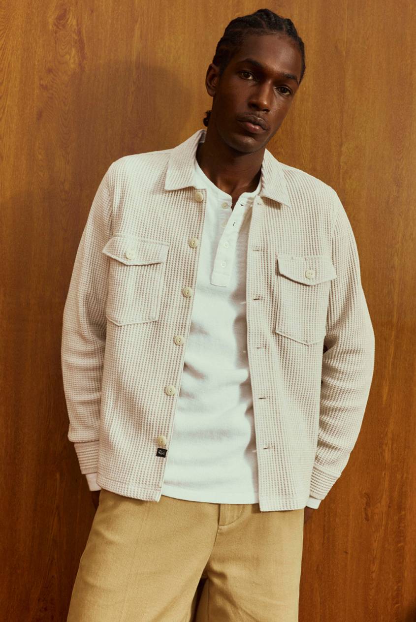 Man standing against a wood-paneled wall, wearing a cream waffle-textured overshirt with chest pockets over a white henley, and khaki pants.