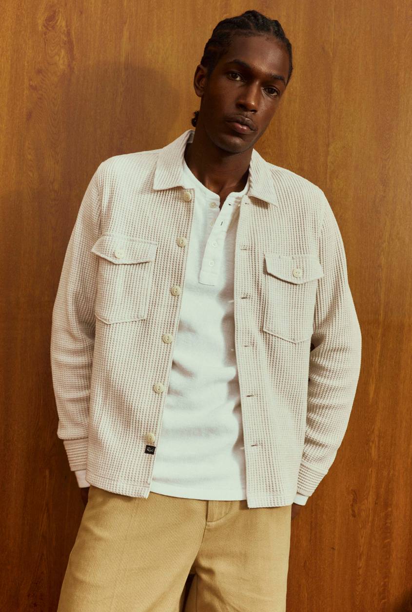 Man with braided hair standing against a wood-paneled wall, wearing a cream waffle-textured overshirt with chest pockets over a white henley, and khaki pants.