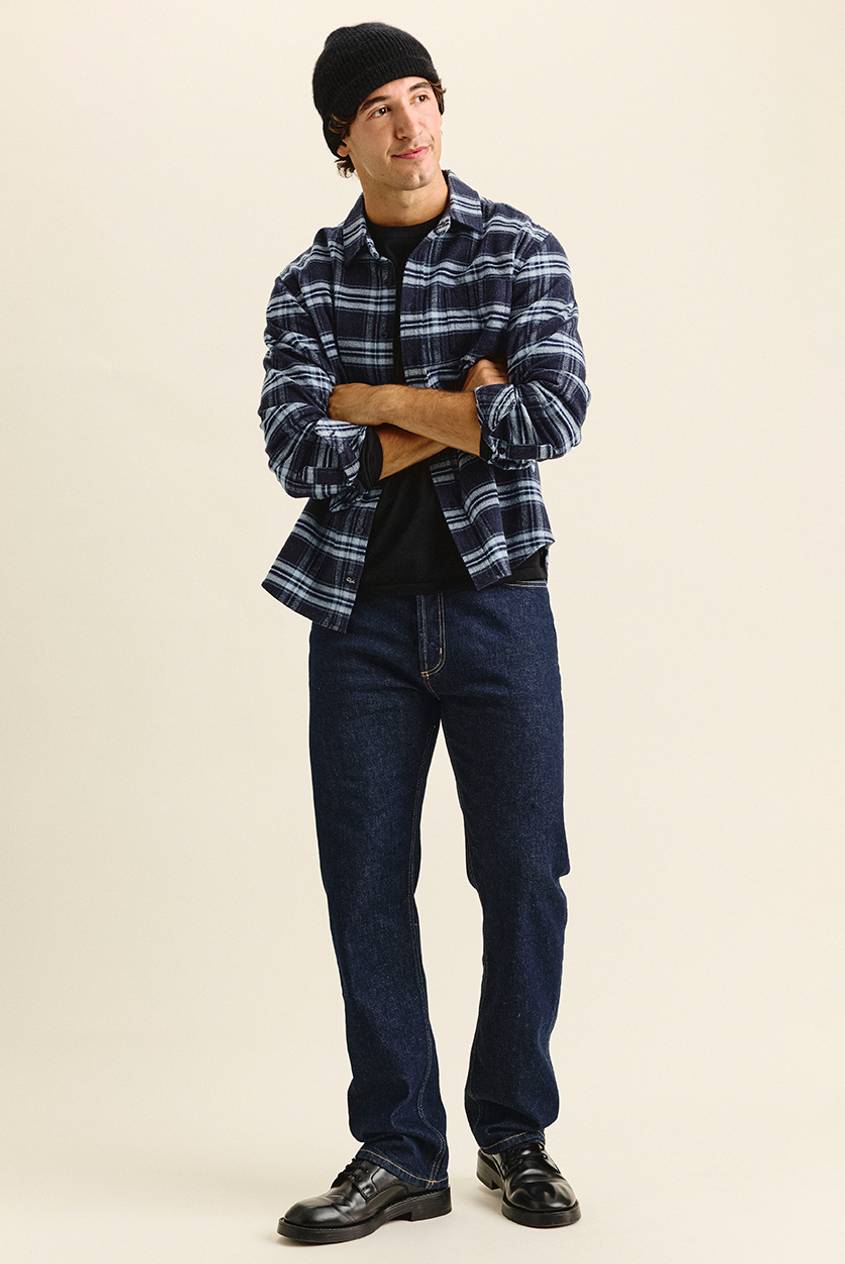 Man standing with arms crossed, looking off to the side, wearing a black knit beanie, a navy and light blue plaid flannel shirt over a black tee, dark indigo jeans, and black lace-up shoes.