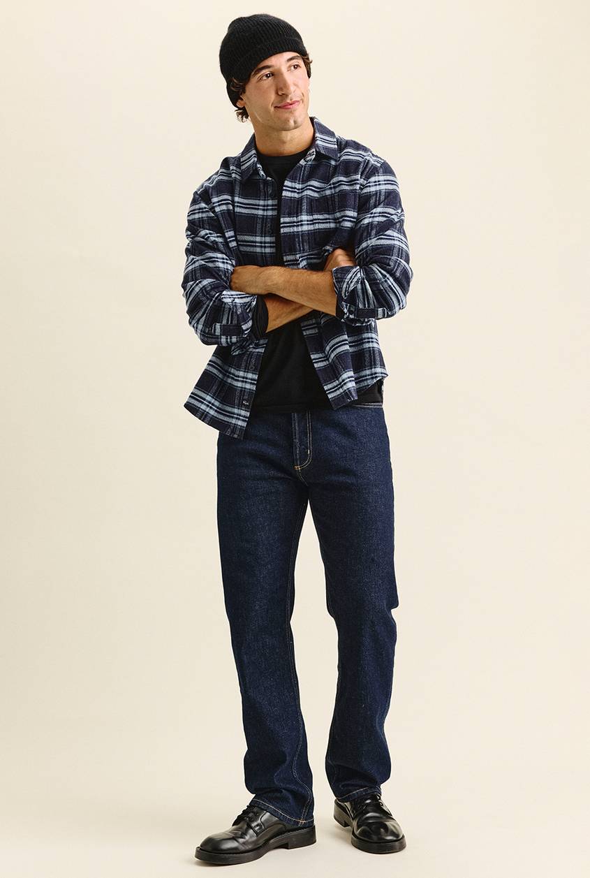 Man standing with arms crossed, looking off to the side, wearing a black knit beanie, a navy and light blue plaid flannel shirt over a black tee, dark indigo jeans, and black lace-up shoes.