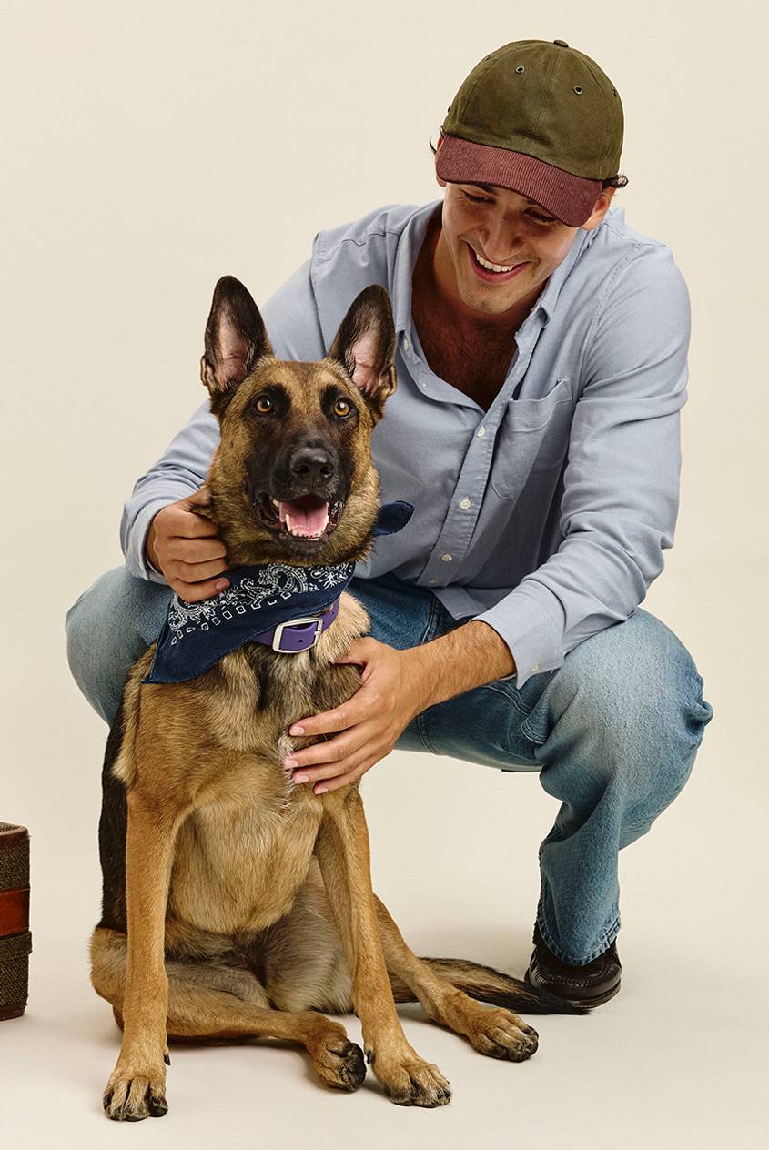 Smiling man crouching beside a German Shepherd, wearing an olive and burgundy baseball cap, a light blue button-up shirt, and light-wash jeans. The dog wears a navy paisley bandana and a purple collar.