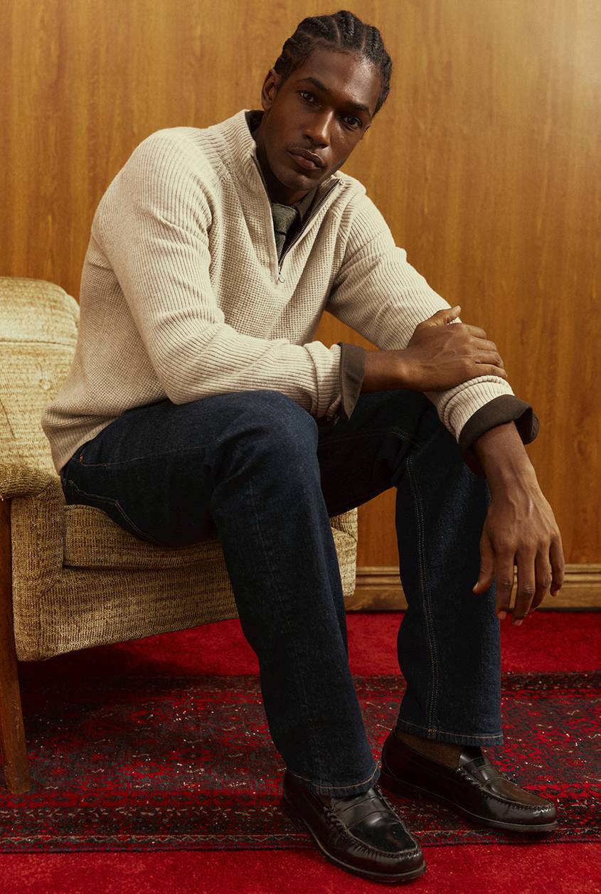 Man with braided hair seated in a tan upholstered chair, wearing a cream waffle-knit quarter-zip sweater over a collared shirt, dark-wash jeans, and burgundy penny loafers. He sits on a red patterned rug against a wood-paneled wall.