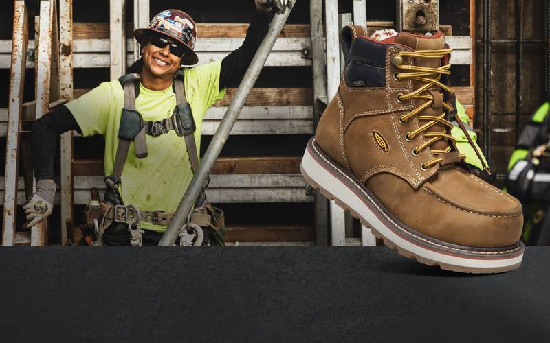 Woman wearing construction-safety gear posing in front of metal building structure with a product shot of the brown, Keen Cincinnati workboot overlayed in front. 