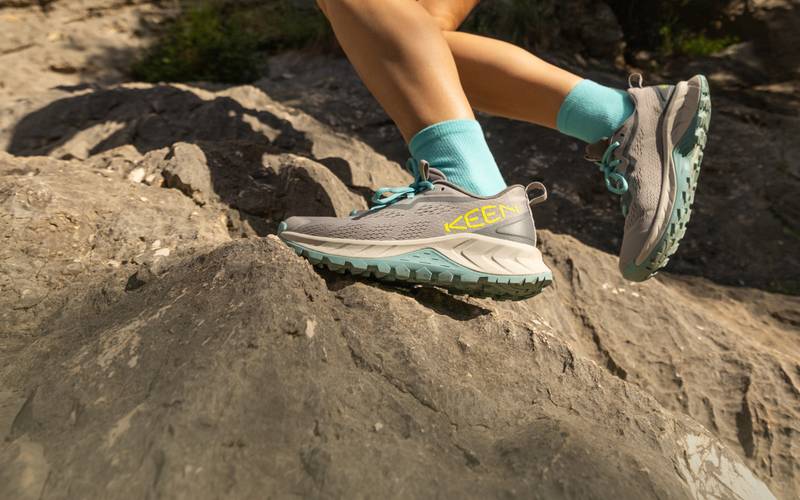 Knee-down shot of woman wearing gray and light blue Versacore speed hiking shoes and running over rocky terrain. 