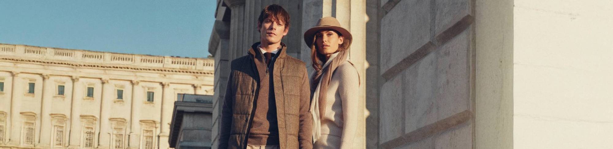 Man and woman standing side by side in sunlight against a stone building; the man wears a brown gilet over a sweater with beige trousers, and the woman wears a light sweater and skirt with a wide-brimmed hat