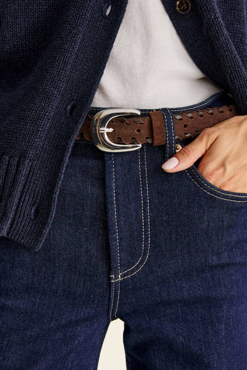 Close-up of a brown suede woven belt with a polished silver buckle, worn with dark indigo jeans and a navy knit cardigan over a white top, one hand in the pocket.