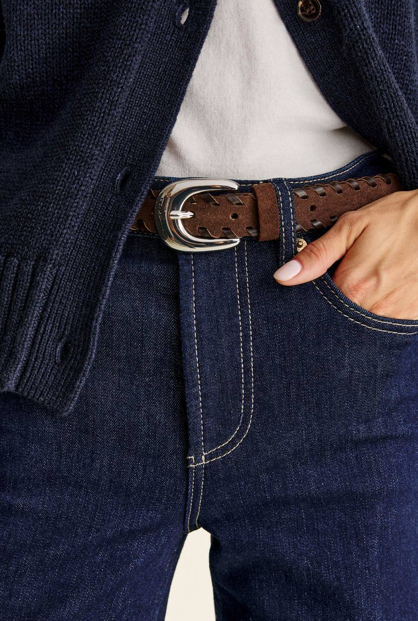 Close-up of a brown suede woven belt with a polished silver buckle, worn with dark indigo jeans and a navy knit cardigan over a white top, one hand in the pocket.