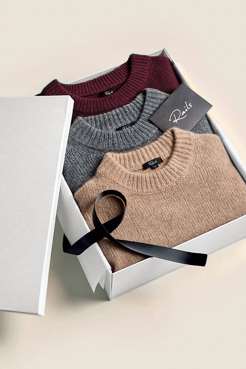 Three folded sweaters in burgundy, gray, and camel sit in an open white gift box with a black satin ribbon and a Rails card, the lid propped to the left.