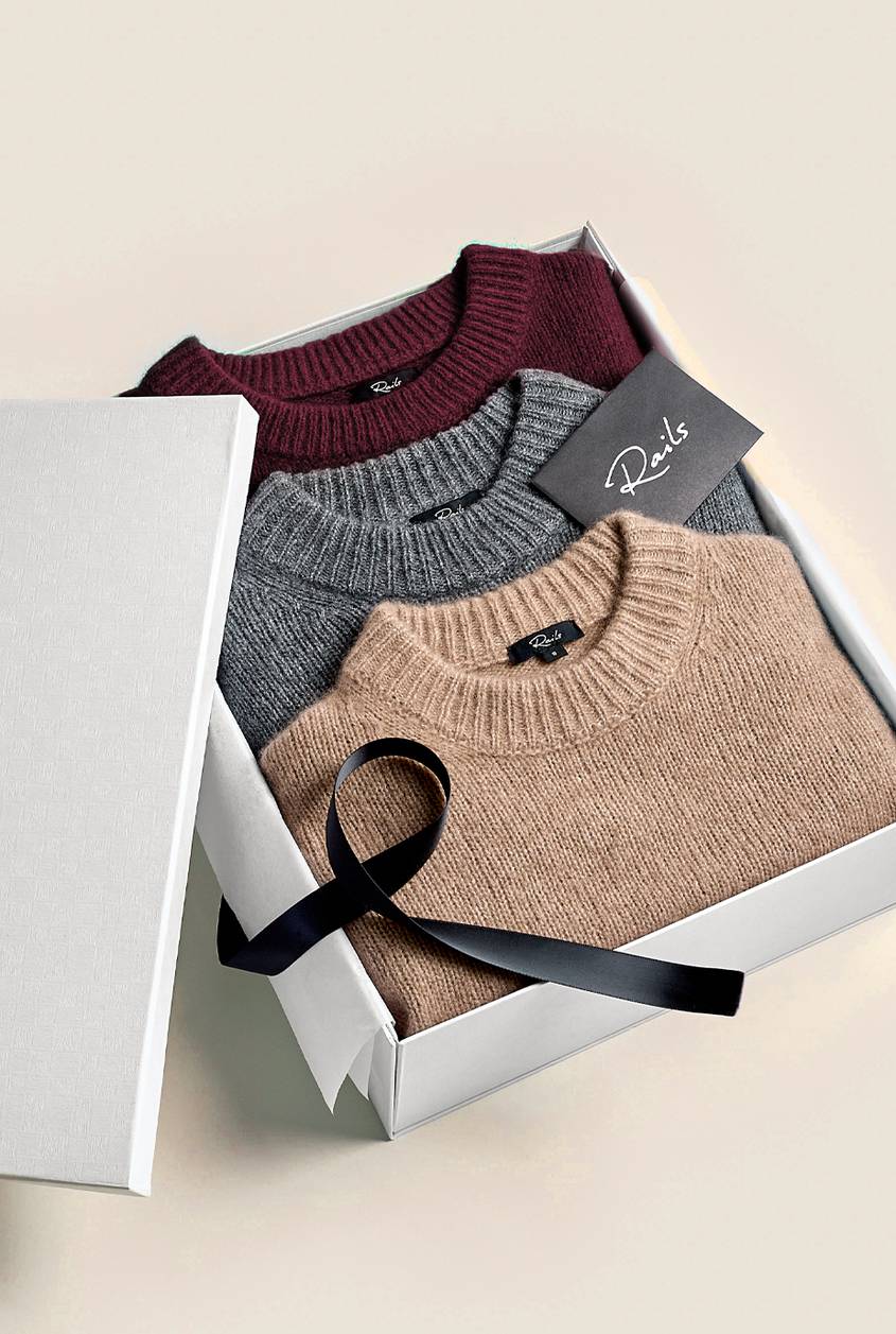 Three folded sweaters in burgundy, gray, and camel sit in an open white gift box with a black satin ribbon and a Rails card, the lid propped to the left.