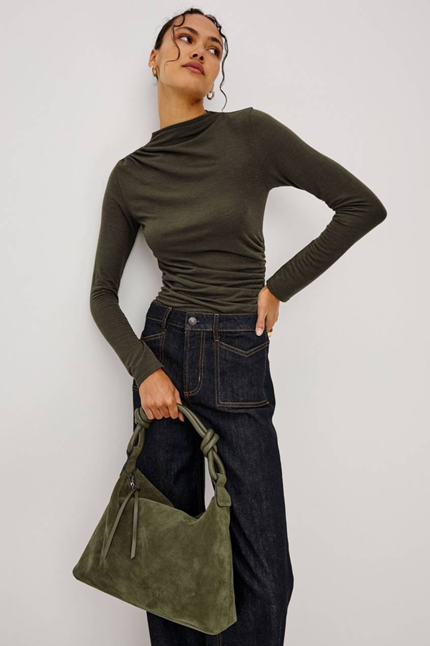 Woman wearing an olive green long-sleeve top and dark jeans, carrying a green suede handbag.