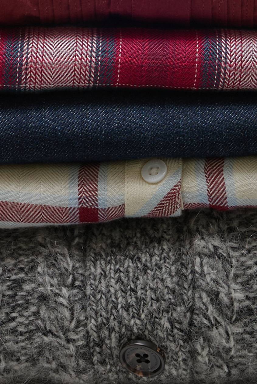Close-up of layered plaid, denim, and textured knit fabrics with button details, showcasing the rich textures and patterns of the fall collection. Close-Up View.