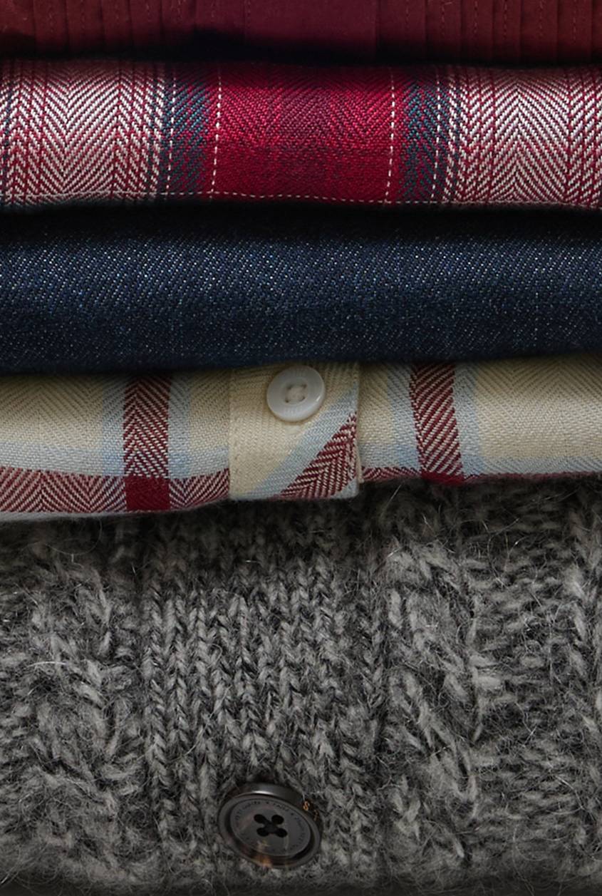 Close-up of layered plaid, denim, and textured knit fabrics with button details, showcasing the rich textures and patterns of the fall collection. Close-Up View.