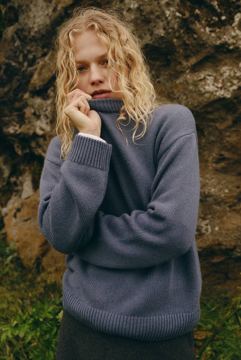 Blonde model wearing an oversized blue knit sweater standing in front of a rocky landscape.