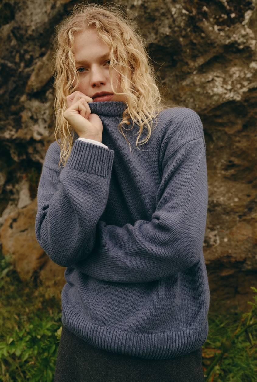 Blonde model wearing an oversized blue knit sweater standing in front of a rocky landscape.