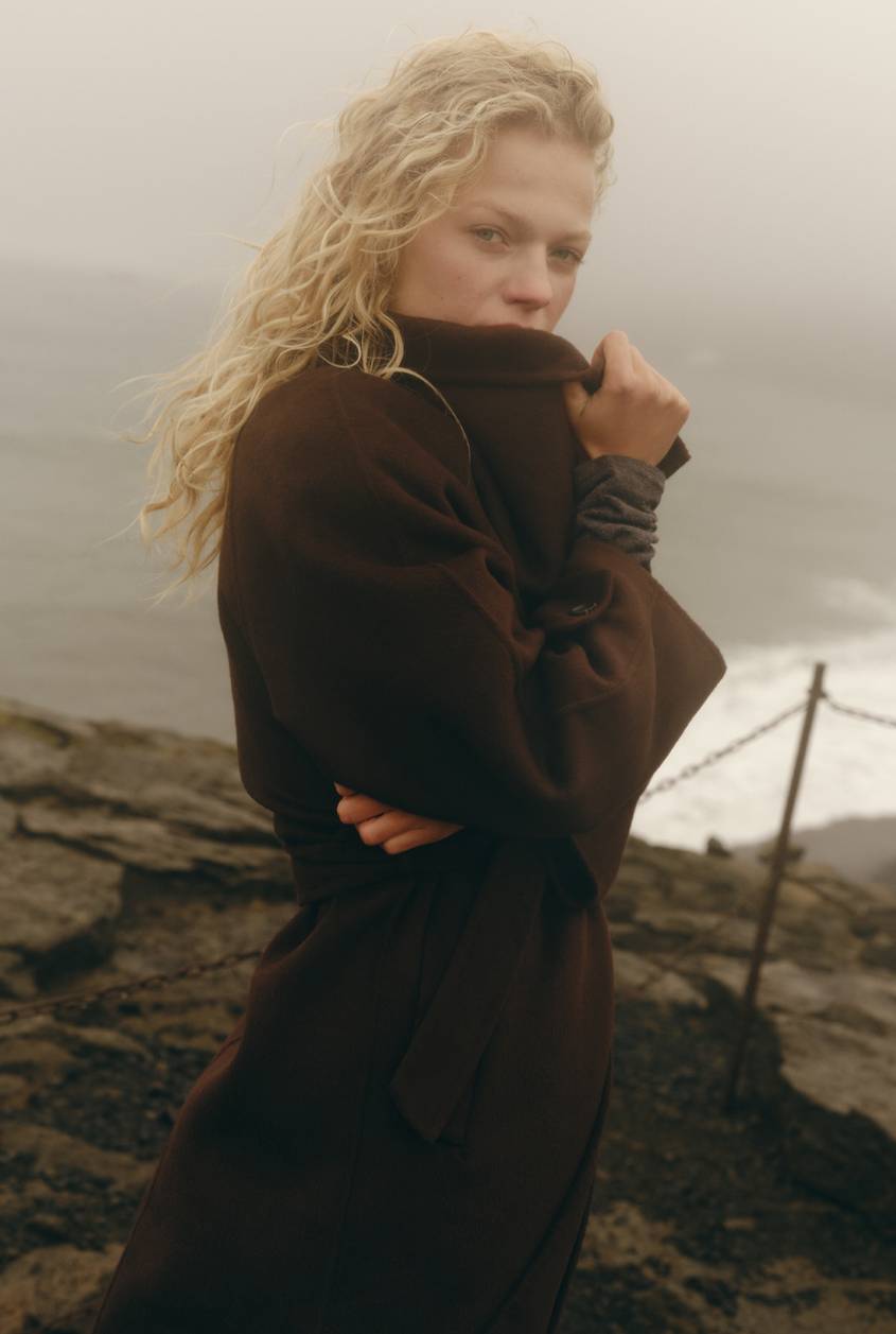 Blonde model wearing a dark brown coat standing on a rocky coastline with the ocean in the background.