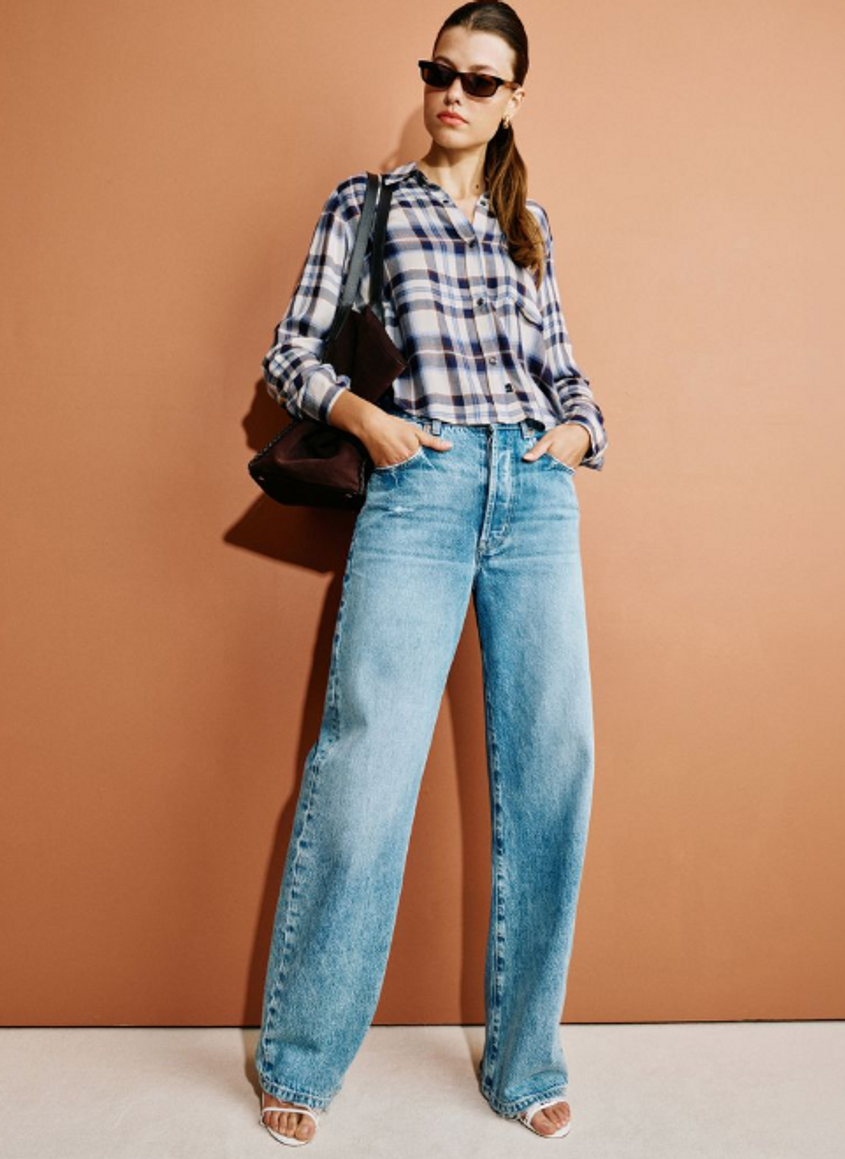 Woman wearing a plaid shirt with blue jeans and accessories while posing against an orange background.