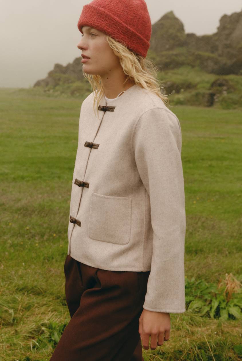 Blonde model wearing a red beanie, cream toggle-front jacket, and brown pants standing in a grassy landscape.