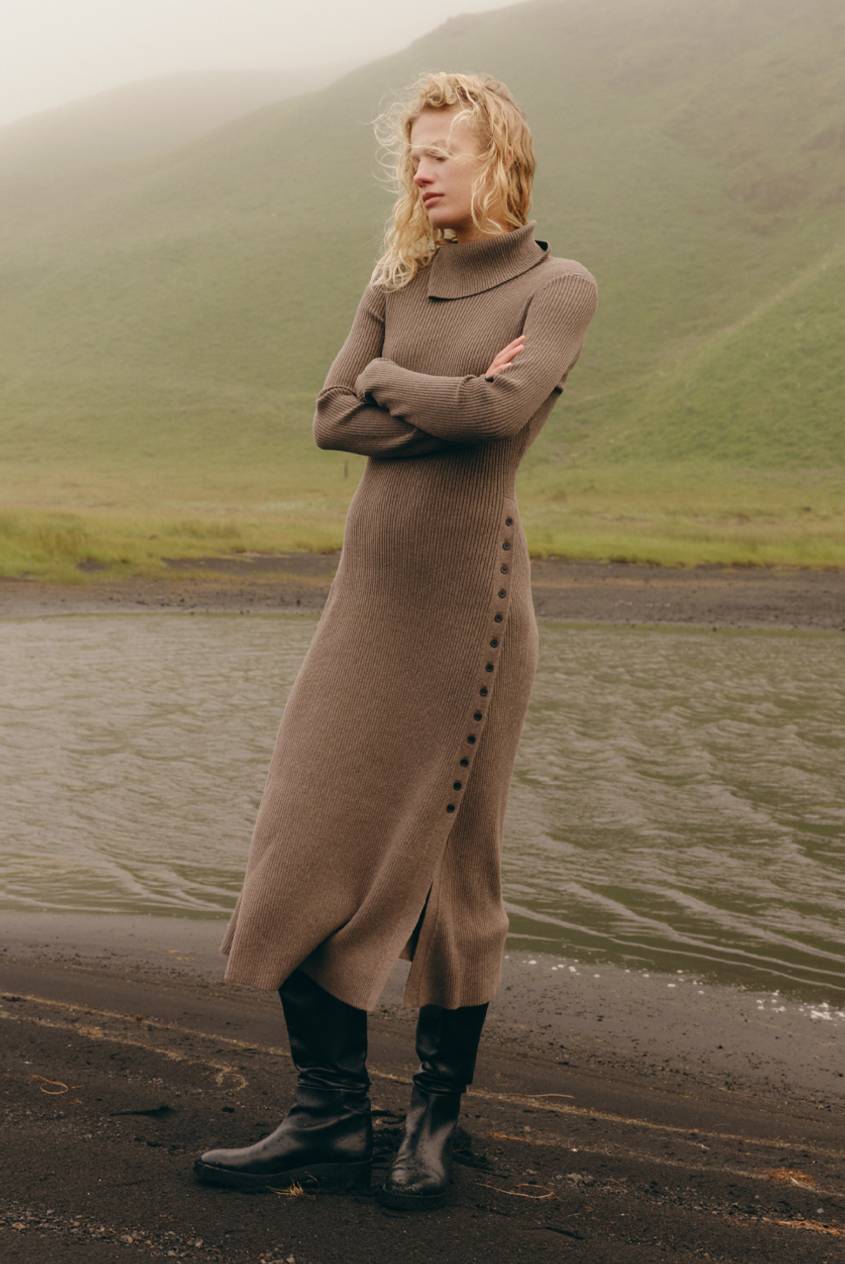 Model in a taupe ribbed turtleneck sweater dress with a side button placket and black knee-high boots, standing on a dark sand beach with arms crossed against a foggy green hillside backdrop.