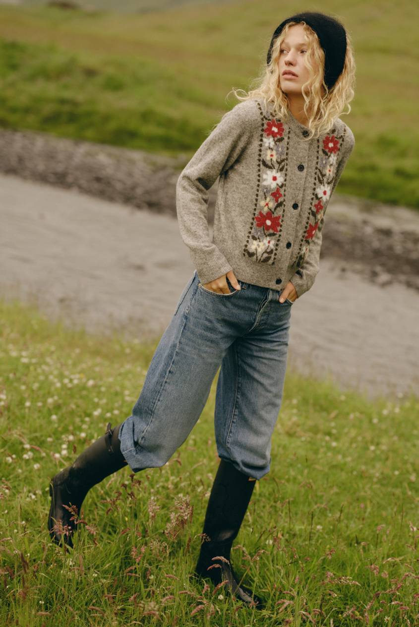 Model wearing a gray floral-embroidered cardigan with red and white flowers, a black beanie, wide-leg light-wash jeans, and black tall boots, walking through a grassy field