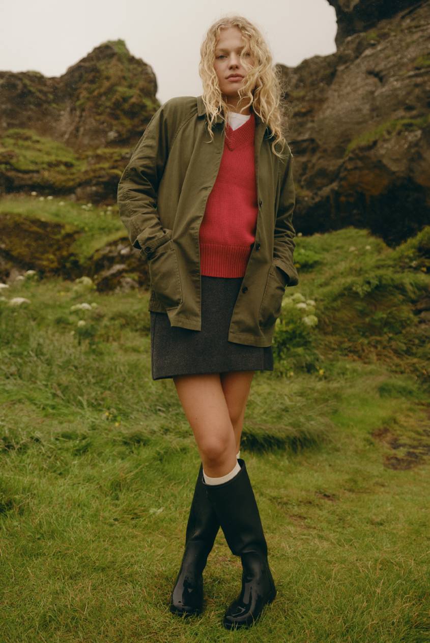 Model wearing an olive utility jacket over a coral V-neck sweater, a charcoal gray mini skirt, cream socks, and black tall rain boots, standing in front of mossy rock formations.