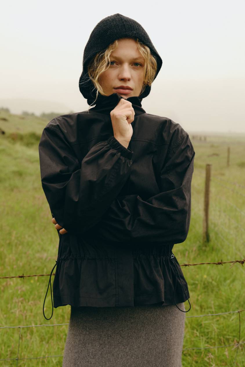 Model wearing a black hooded jacket and gray skirt standing in a grassy field with a fence in the background.