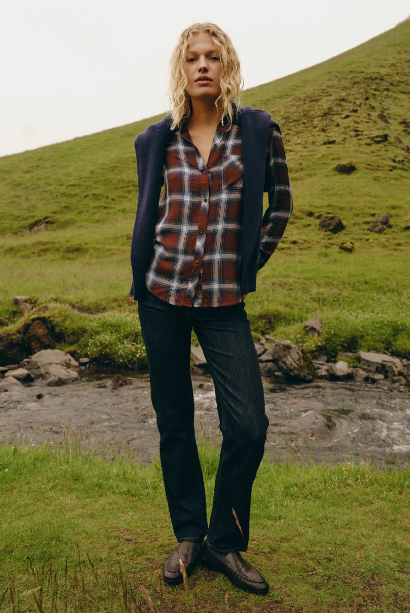 Model wearing a rust and navy plaid button-up shirt with a navy cardigan draped over the shoulders, dark straight-leg jeans, and brown leather loafers, standing on a grassy hillside next to a stream