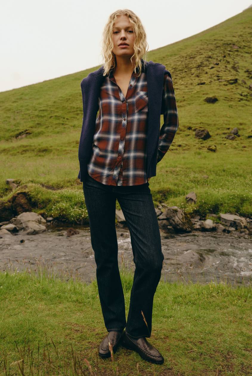 Model wearing a rust and navy plaid button-up shirt with a navy cardigan draped over the shoulders, dark straight-leg jeans, and brown leather loafers, standing on a grassy hillside next to a stream