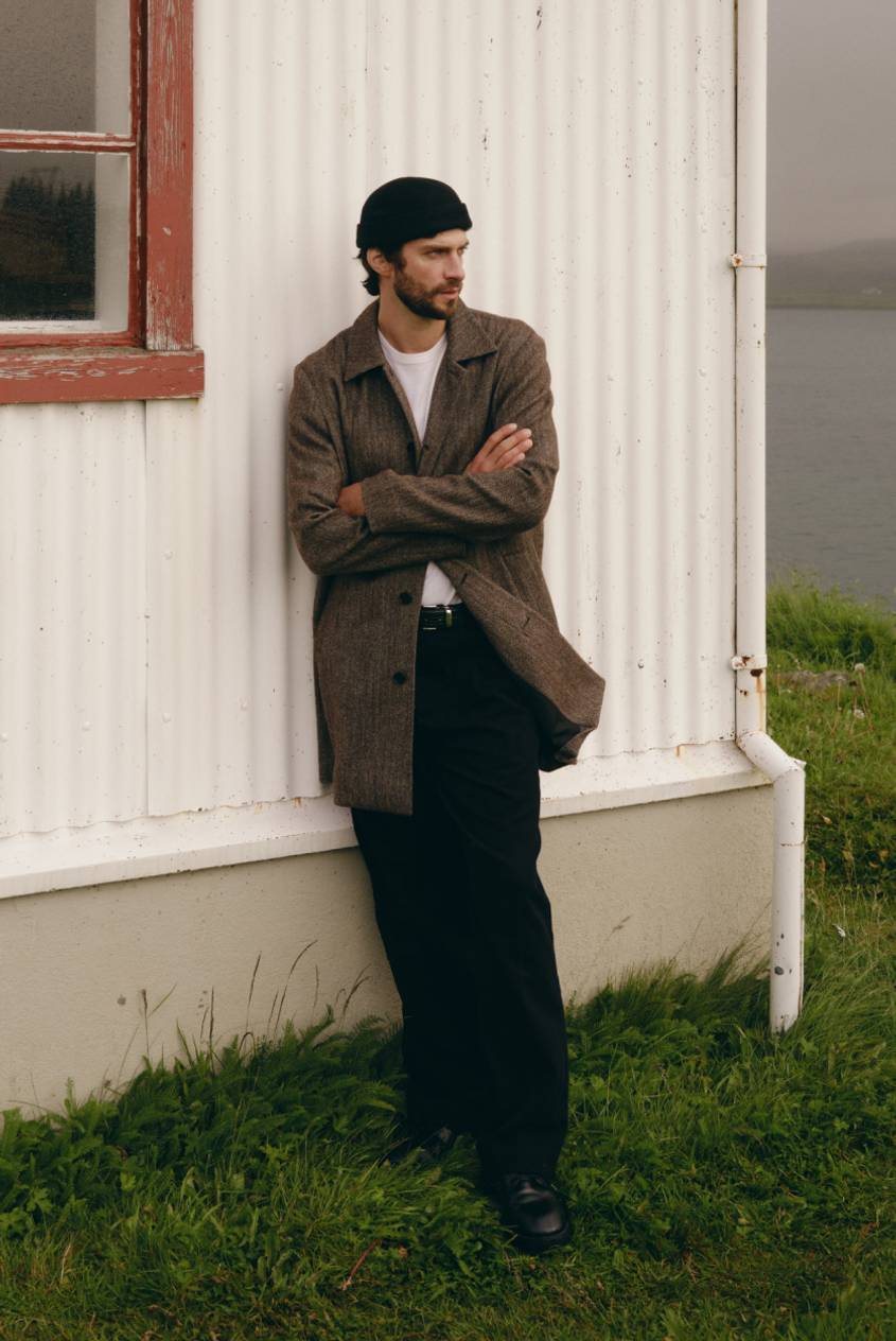 Model wearing a brown wool-blend jacket over a white T-shirt with black pants and loafers, standing outdoors.
