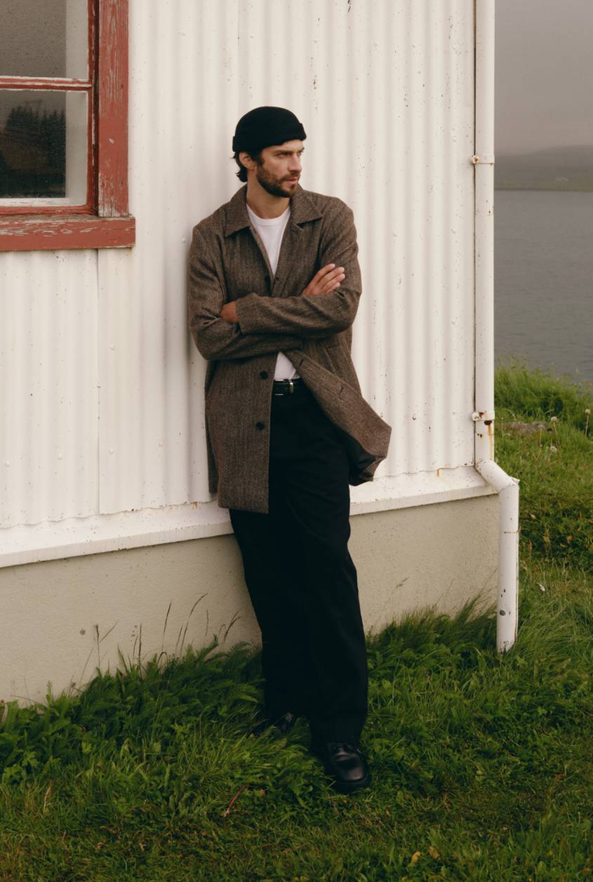 Model wearing a brown wool-blend jacket over a white T-shirt with black pants and loafers, standing outdoors.