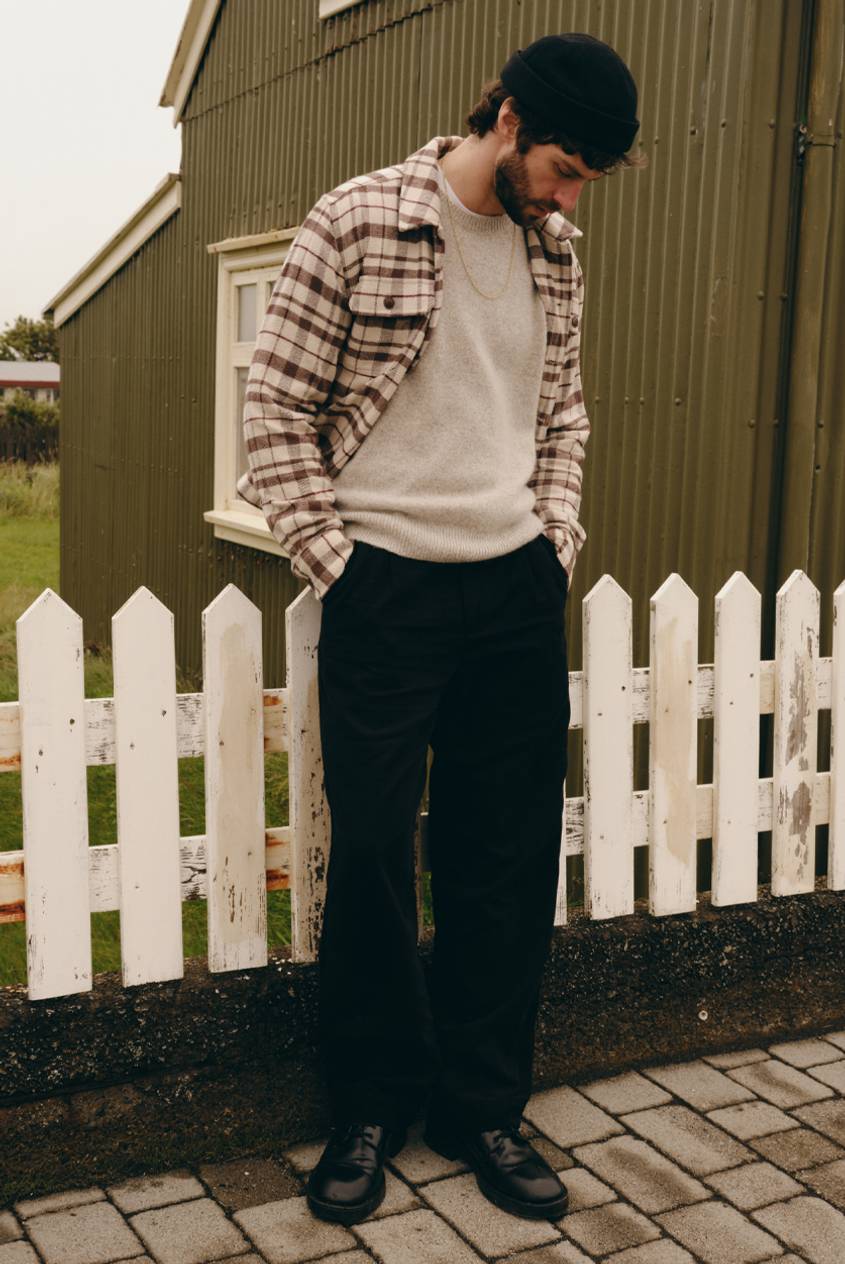Model wearing a brown and cream plaid overshirt over a beige sweater with black pants, standing outdoors.