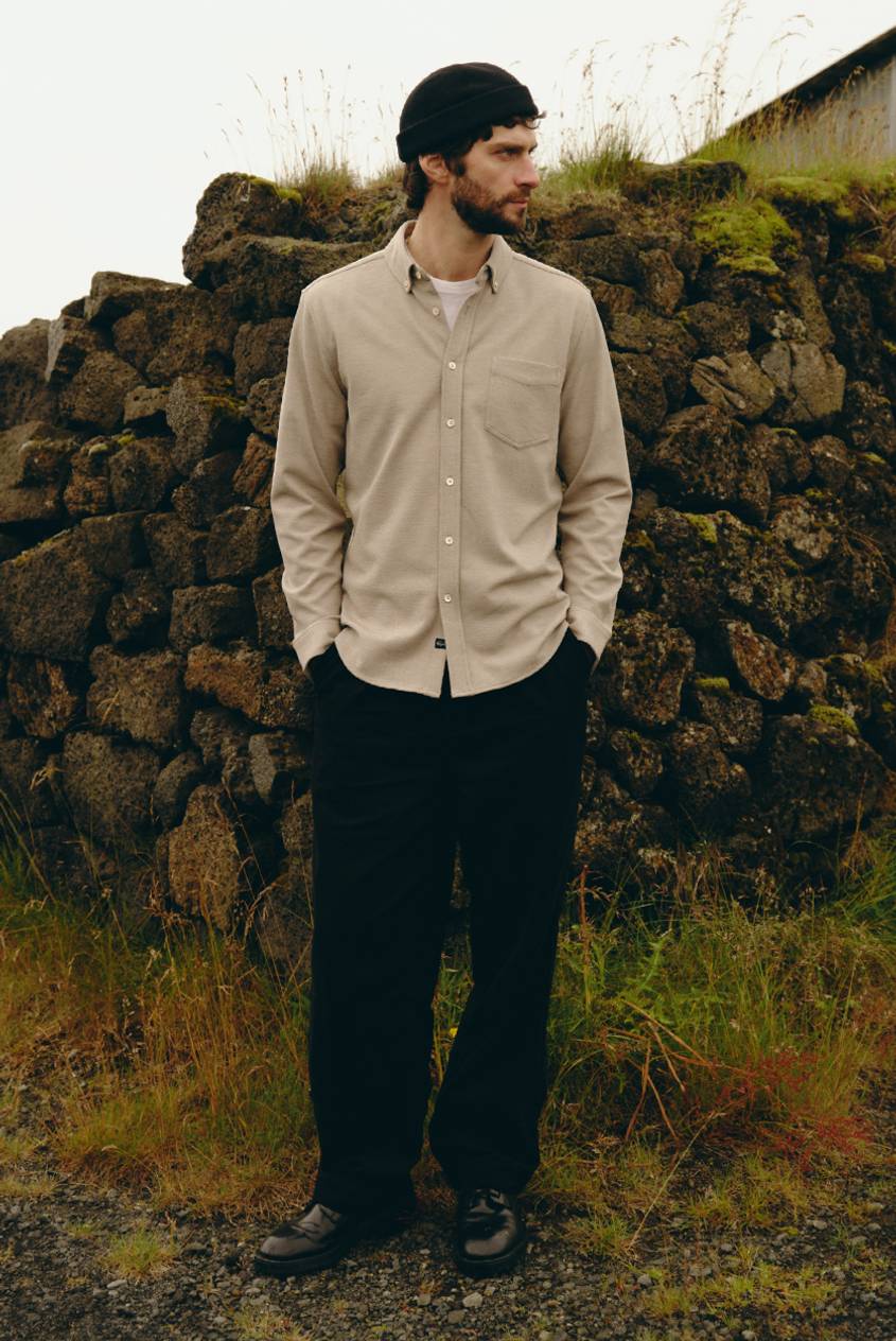 Model wearing a beige long-sleeve button-down shirt with a chest pocket and black pants, standing outdoors.
