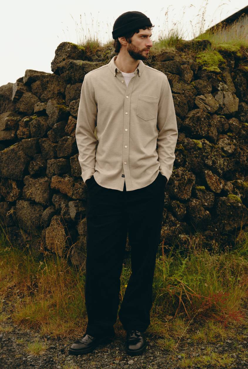 Model wearing a beige long-sleeve button-down shirt with a chest pocket and black pants, standing outdoors.