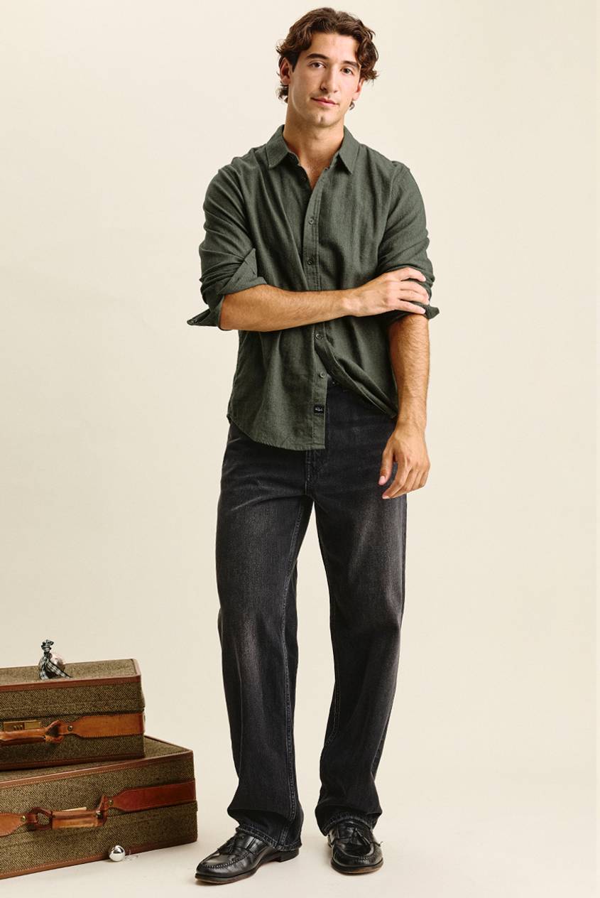 Man with wavy brown hair standing with one hand rolling up his sleeve, wearing an olive green button-up shirt, wide-leg faded black jeans, and black tassel loafers. Vintage tweed suitcases with leather straps are stacked beside him.