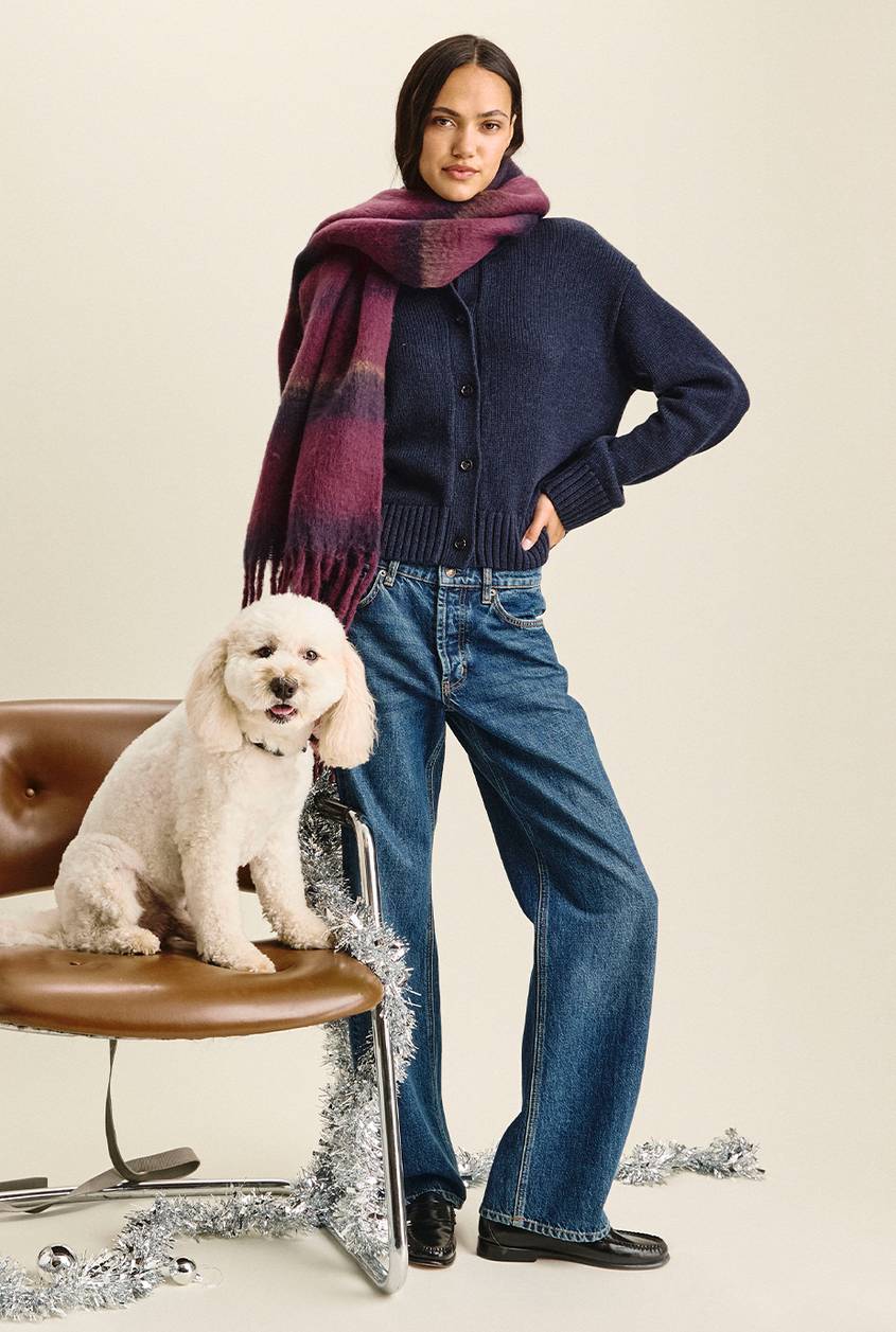 Woman standing with one hand on her hip, wearing a navy button-front cardigan, a plum and navy fringed scarf, wide-leg dark-wash jeans, and black loafers. A white doodle dog sits on a brown leather chair beside her, with silver tinsel draped around the chair.